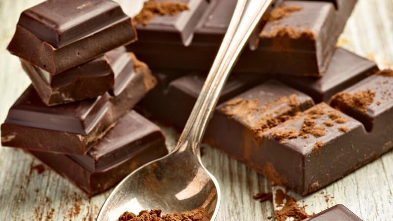 Silberner Löffel mit Kakaopulver liegt auf Tafelschokolade auf weiß-grauem Holztisch