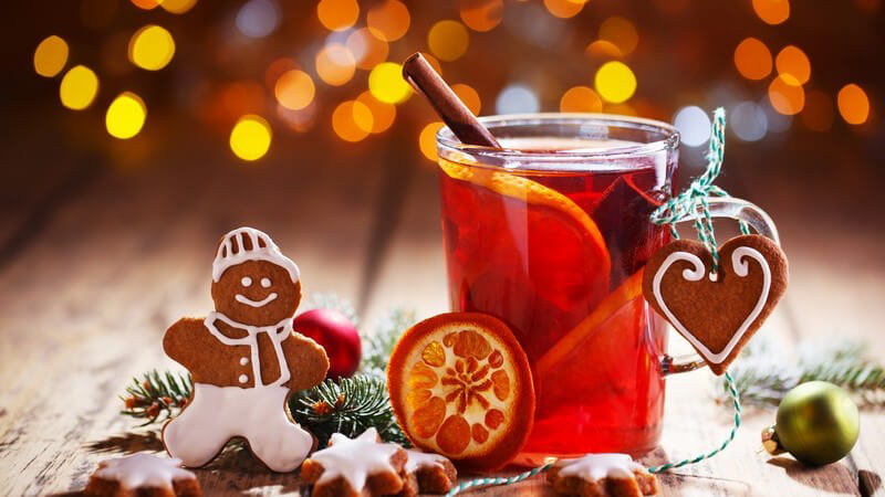 Glühweinglas mit Orangenscheiben und Zimtstange neben Weihnachtsplätzchen und -dekoration auf Holztisch