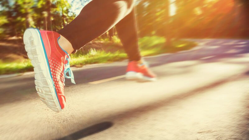 Joggerin in roten Laufschuhen und schwarzer Laufhose läuft im Sonnenschein eine Straße entlang