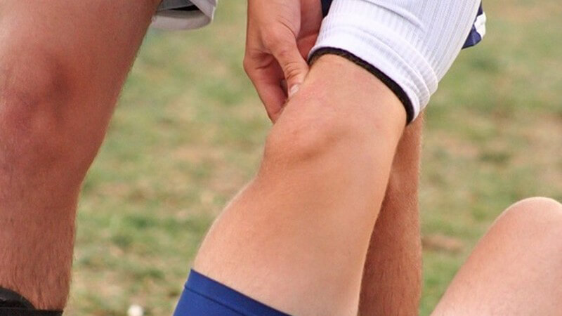 Fußballspieler richtet verletztes Bein eines am Boden liegenden anderen Spielers, hält Wade, keine Gesichter sichtbar