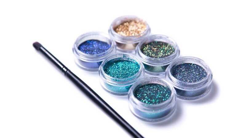 Sechs Gläschen mit Glitzer-Make-up neben Pinsel auf weißem Untergrund