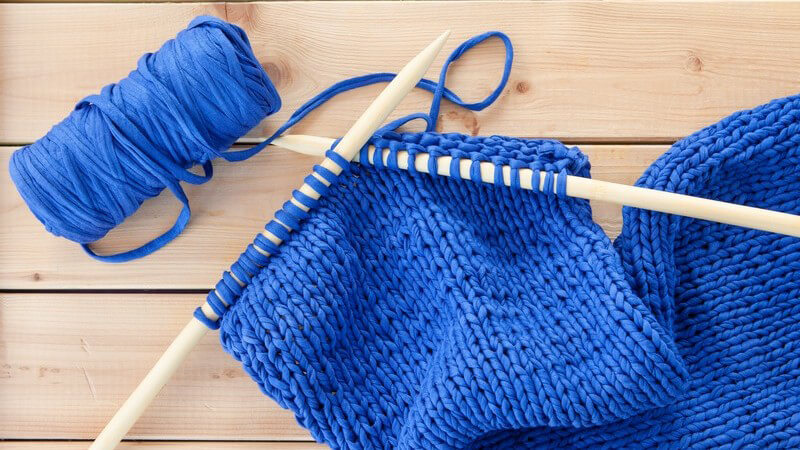 Zwei hölzerne Stricknadeln mit blauem Textilgarn auf hellem Holzuntergrund
