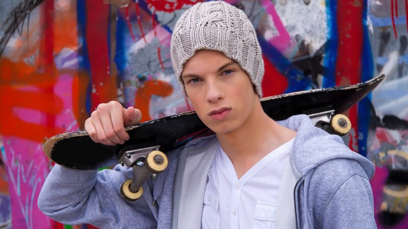 Junge mit grauer Strickmütze trägt Skateboard auf Schulter