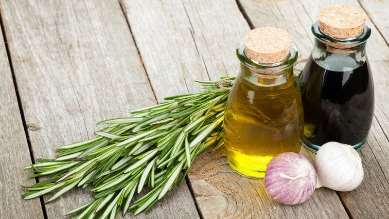 Korkverschluss-Fläschchen mit Öl und Essig neben Knoblauch und Rosmarin auf Holz