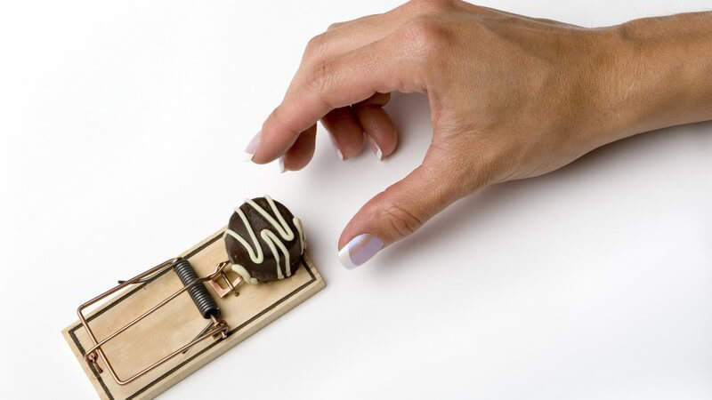 Frauenhand greift nach Praline, die in Mausefalle liegt, weißer Hintergrund