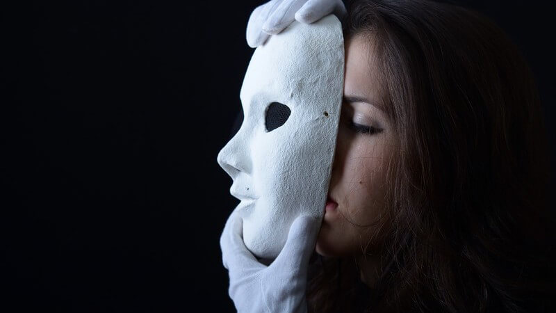 Frau mit weißen Handschuhen hält sich eine weiße Theatermaske vor das Gesicht, schwarzer Hintergrund