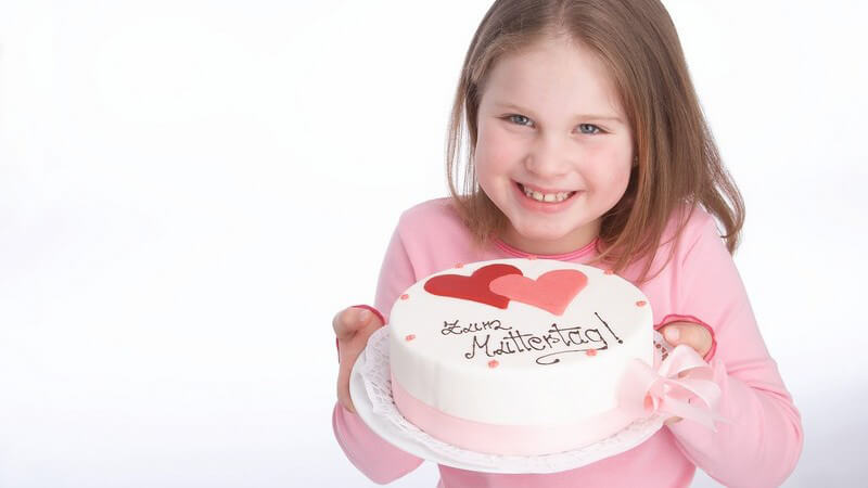 Kleines lachendes Mädchen hält Torte mit Herzen und Aufschrift "Zum Muttertag" in die Kamera