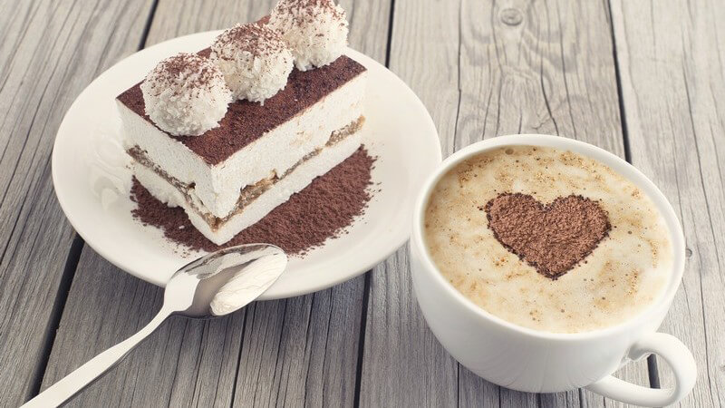 Stück Torte neben Kaffeetasse mit Herzmuster im Schaum auf Holztisch
