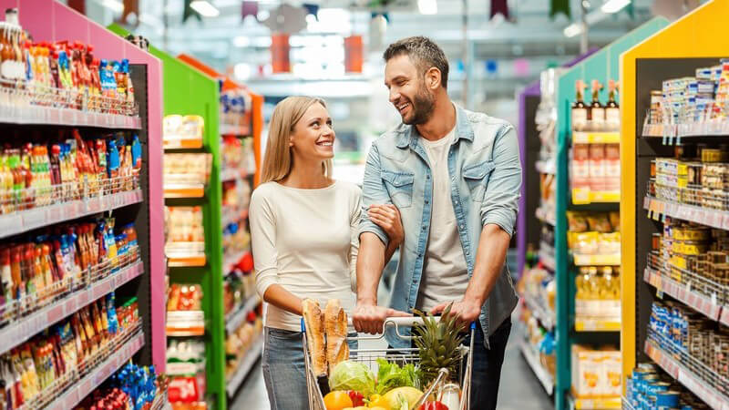 Paar beim gemeinsamen Einkaufen im Supermarkt, schiebt einen vollen Einkaufswagen und sieht sich verliebt an