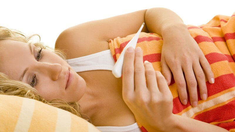 Blonde Frau im Bett schaut auf digitales Fieberthermometer nach Fiebermessen