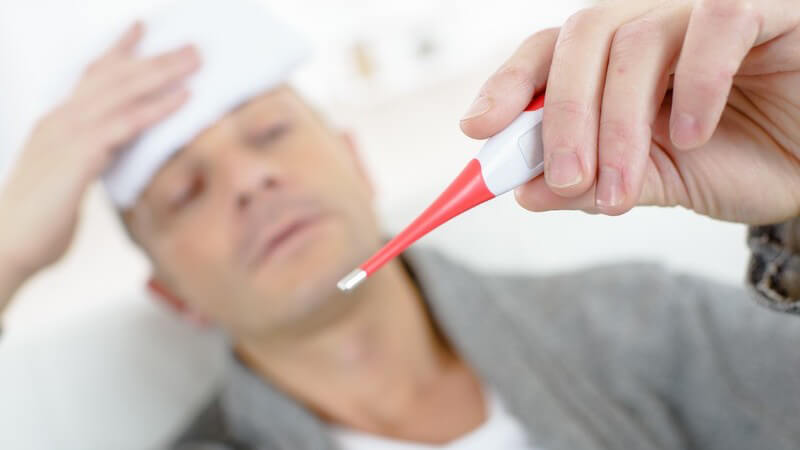Kranker Mann hält sich einen Waschlappen an die Stirn und guckt auf ein rotes Fieberthermometer