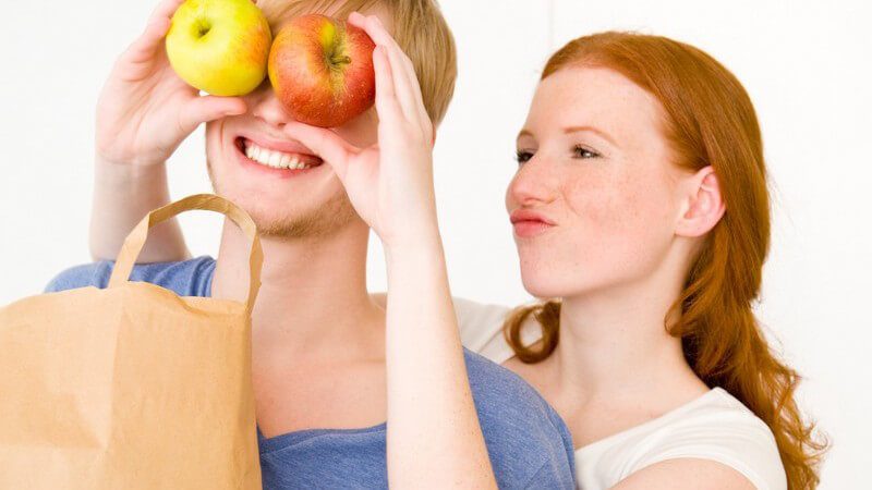 Junges Paar, sie steht hinter ihm und hält ihm Äpfel auf Augen, er trägt Einkaufstüte