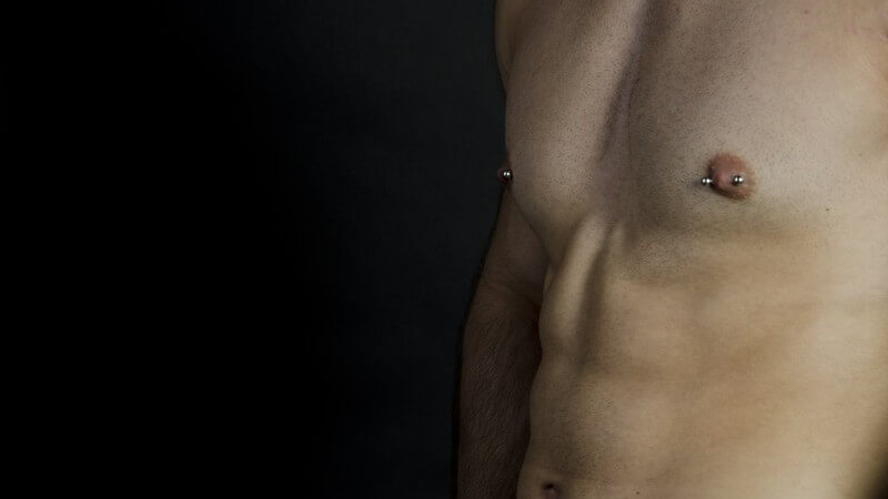 Muskulöser Oberkörper eines Mannes mit Brustwarzenpiercings vor schwarzem Hintergrund