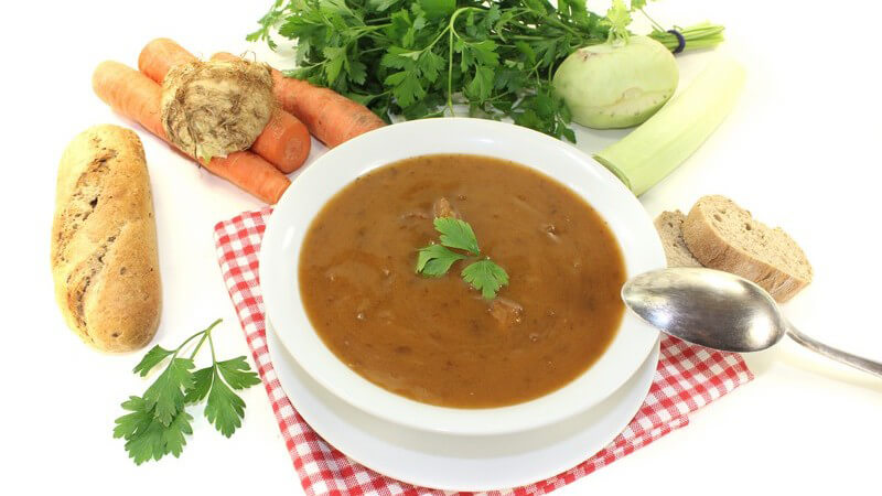 Ochsenschwanzsuppe mit Petersilie, daneben Brot und Suppengemüse vor weißem Hintergrund