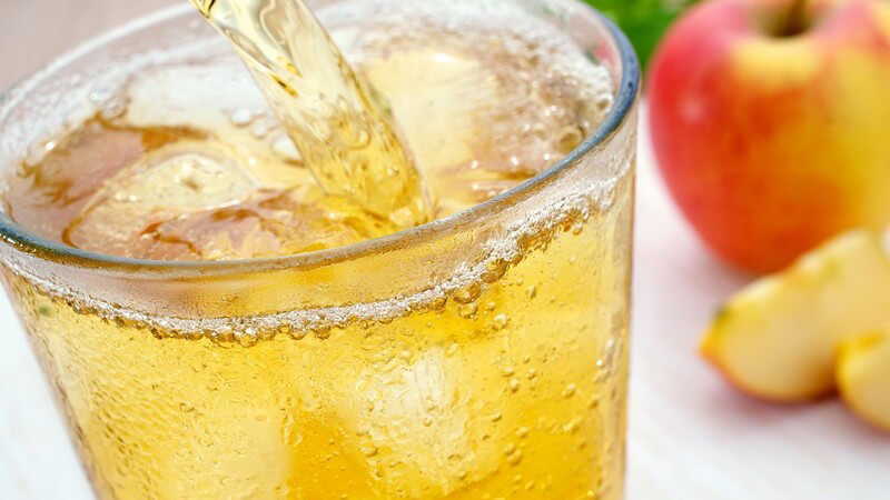 Apfelschorle fließt in Glas mit Eiswürfeln, daneben liegen Äpfel