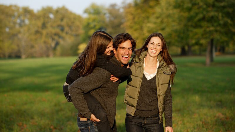 Junge lachende Familie draußen, Vater trägt Tochter auf Rücken