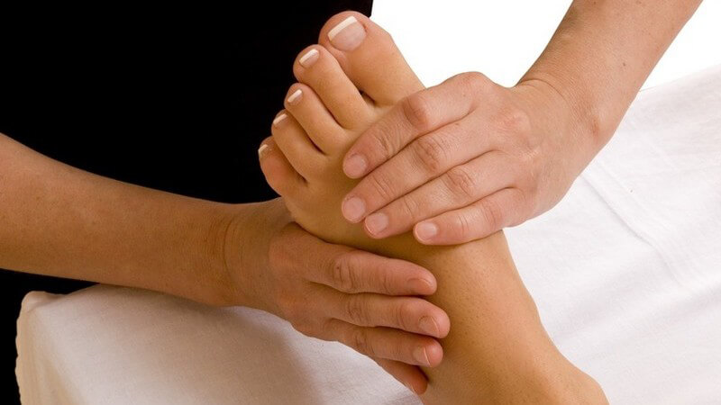 Linker manikürierter Fuß einer Frau liegt auf weißem Laken und wird von zwei Händen einer Frau massiert (Fussmassage