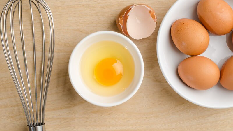 Aufgeschlagenes Ei in Schüsselchen, daneben Schüssel mit braunen Eiern und ein Schneebesen