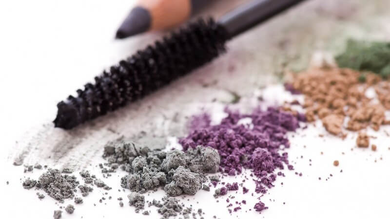 Lidschattenbrösel in grau, lila, orange und grün, dahinter Mascarabürste und Kajalstift