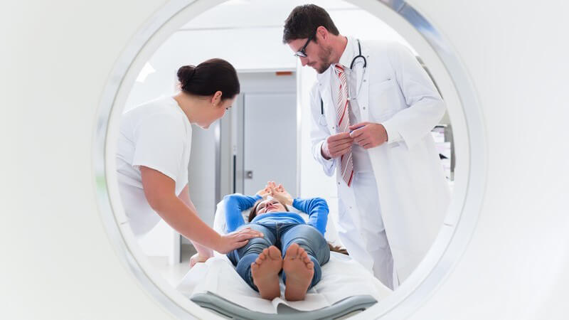 Patientin auf Liege eines Computertomographen, neben ihr stehen Arzt und Krankenschwester