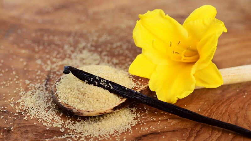 Vanillezucker auf einem Holzlöffel, daneben eine Vanilleschote und -blüte 