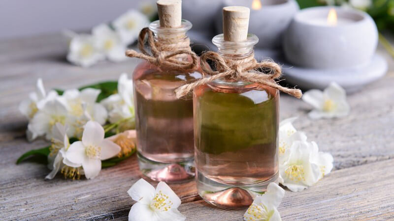 Zwei Aromaölfläschchen umgeben von Jasminblüten auf Holztisch, im Hintergrund brennen drei Kerzen
