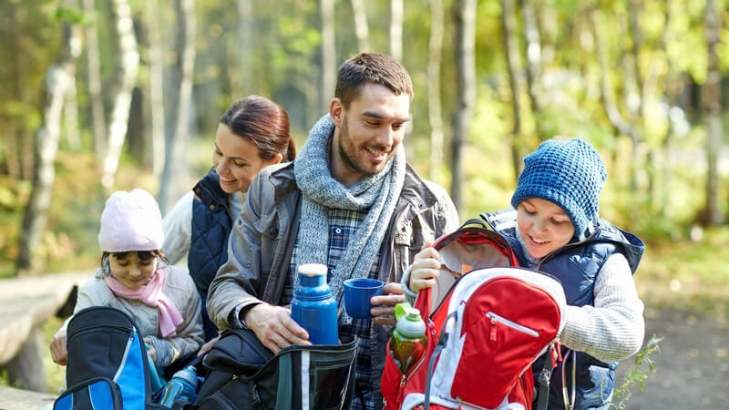Junge Familie mit zwei Kindern sitzt im Wald und macht Picknick, warme Kleidung und Rucksäcke dabei