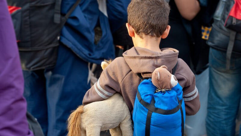 Flüchtlingsjunge steht in einer Gruppe, trägt blauen Schlafsack auf dem Rücken und Kuscheltiere