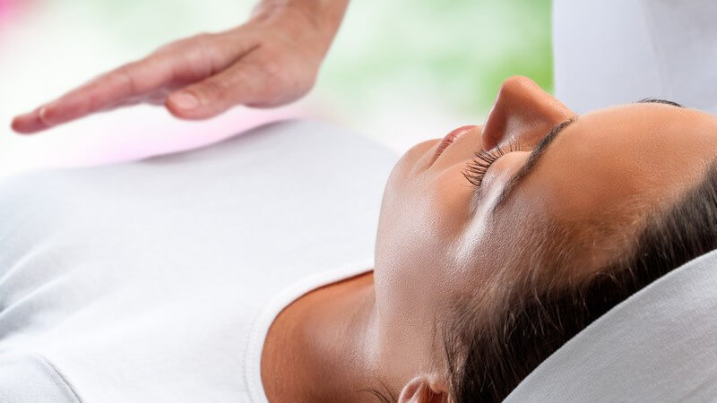 Frau mit weißem Stirnband liegt bei einer Reiki-Behandlung auf dem Rücken, eine Hand schwebt über ihrem Bauch