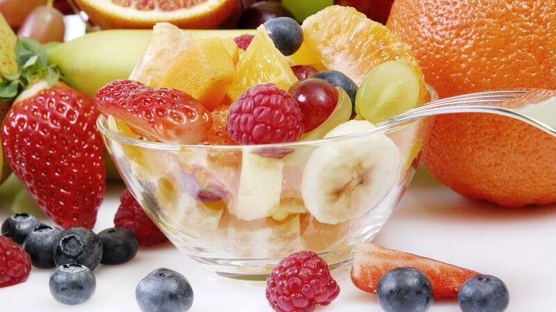 Obstsalat in Glasschüssel, umgeben von frischem Obst auf weißem Hintergrund