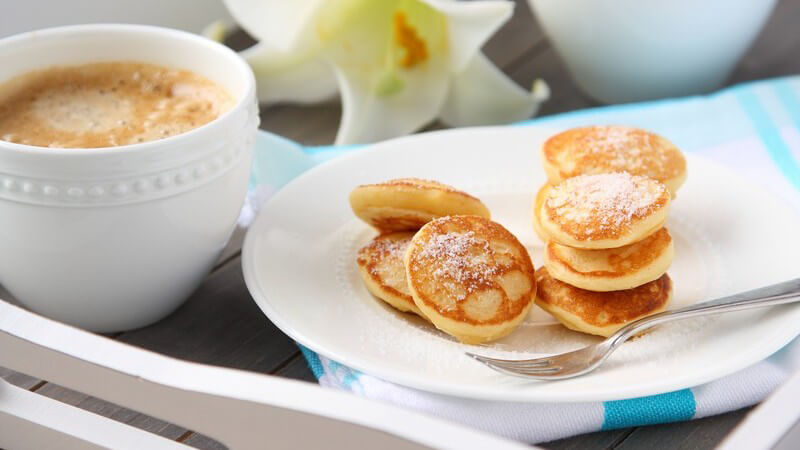 Teller mit niederländischen Mini-Pfannkuchen (Poffertjes) auf einem Tablett neben Tasse Kaffee