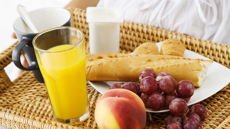 Frau hält Tablett mit Kaffee, Saft, Baguette und Obst auf ihrem Schoß