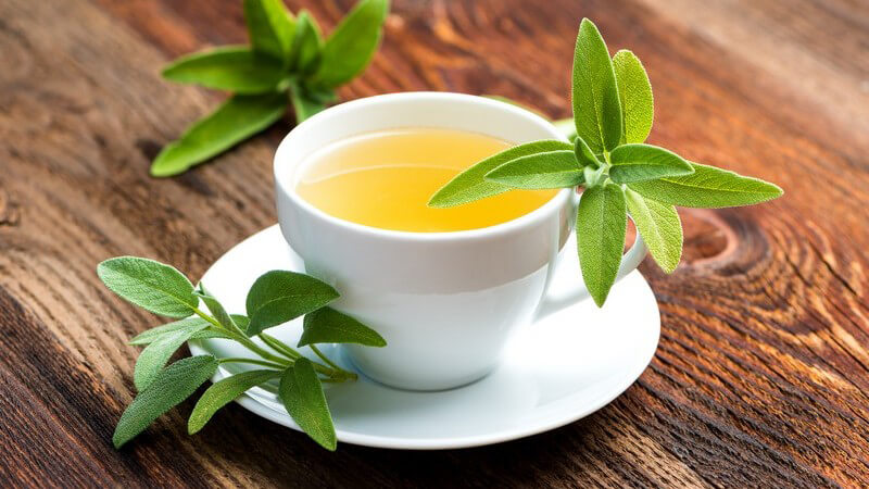 Weiße Tasse mit Salbeitee auf Holztisch, verziert mit Salbeiblättern