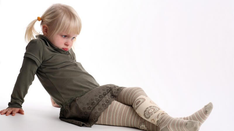 Blondes Mädchen mit Zöpfen und grünem Kleid sitzt auf dem Boden und guckt traurig nach unten