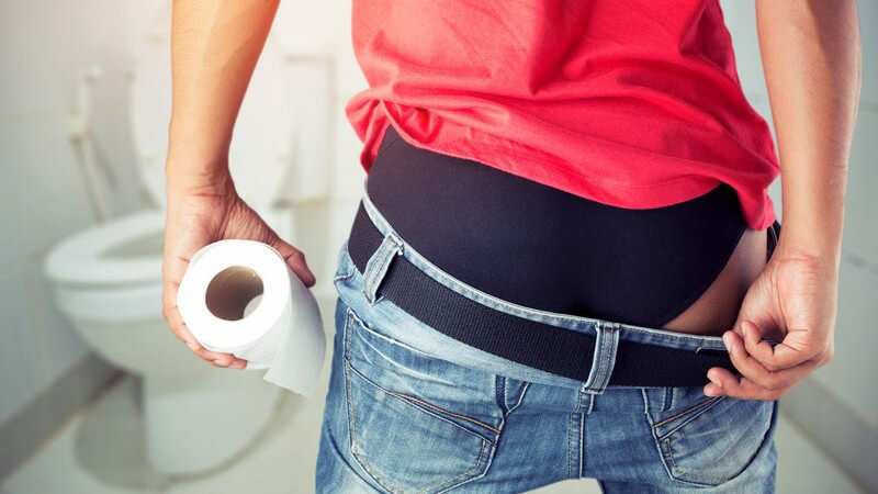 Mann in rotem Shirt auf dem Weg zur Toilette, die Jeans bereits über den Po gezogen und eine Rolle Klopapier in der Han