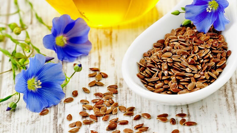 Leinsamen in Schüssel, dahinter Leinöl und blaue Blüten