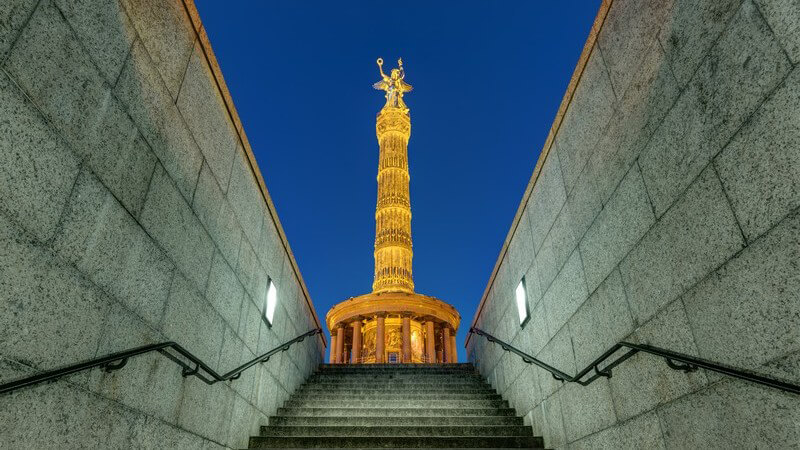 Blick zwischen zwei Wänden einer langen Treppe hindurch auf die Siegessäule in Berlin bei Dämmerung