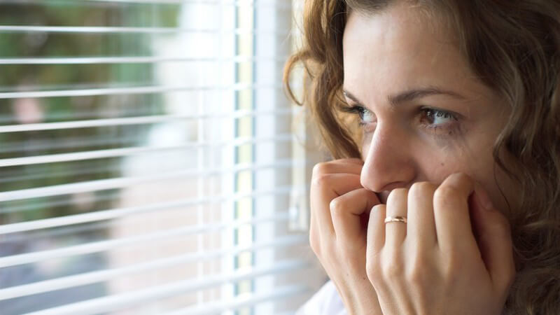 Junge ängstliche Frau mit verweinten Augen steht vor Fenster und hält sich Hände vor Mund