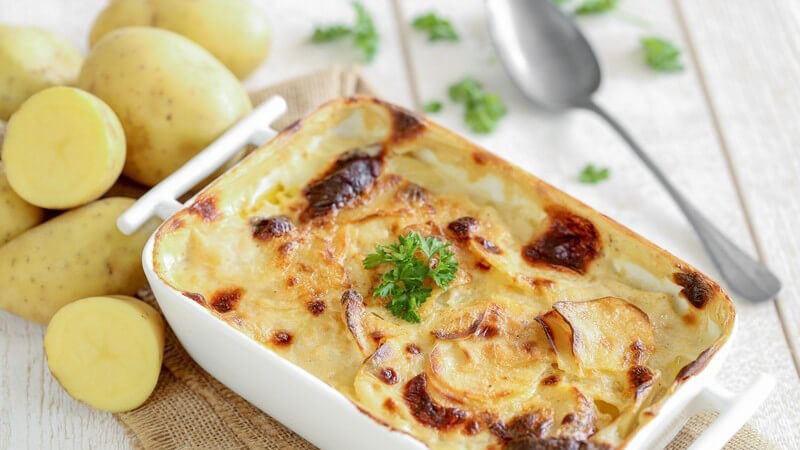 Kartoffelgratin in kleiner weißer Auflaufform neben rohen Kartoffeln und Löffel