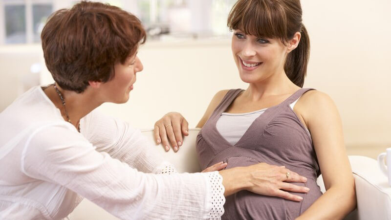 Schwangere Frau sitzt auf einem weißen Sofa und wird von einer Hebamme umsorgt