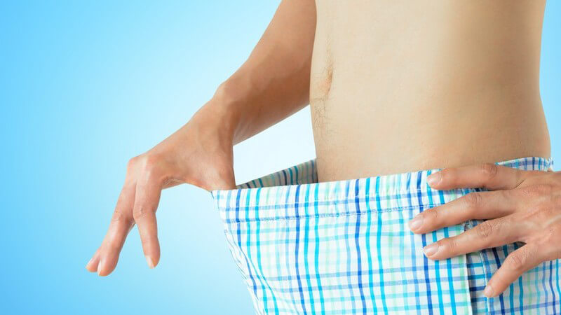 Junger Mann mit nacktem Bauch wirft einen Blick in seine blau gestreifte Boxershorts, hellblauer Hintergrund