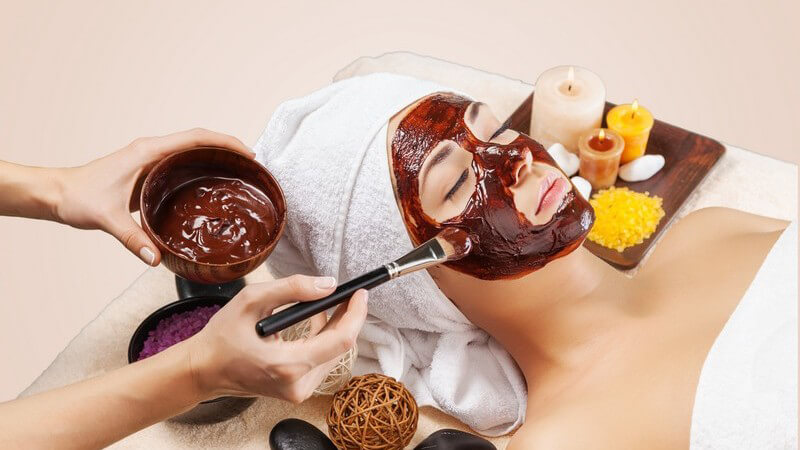 Frau liegt mit Handtuch im Haar im Kosmetikstudio und bekommt eine Schokoladen-Gesichtsmaske aufgepinselt