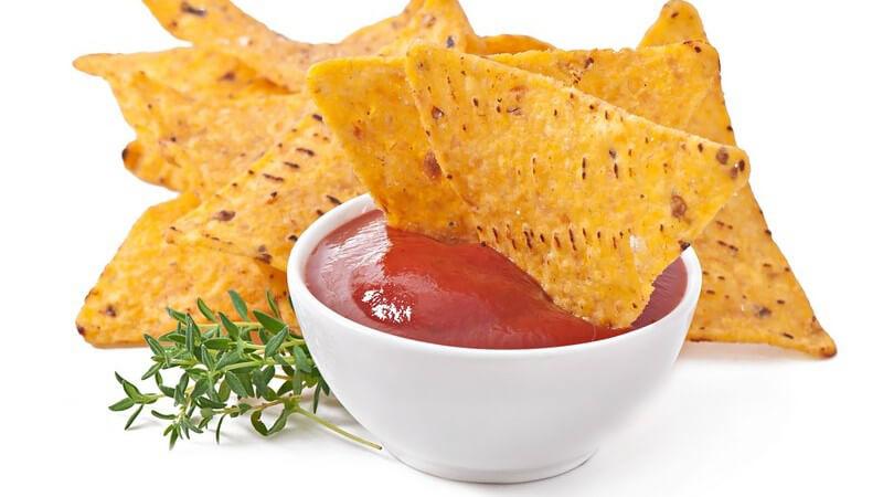 Roter Dip mit zwei Nachos, daneben weitere Nachos und Thymian vor weißem Hintergrund