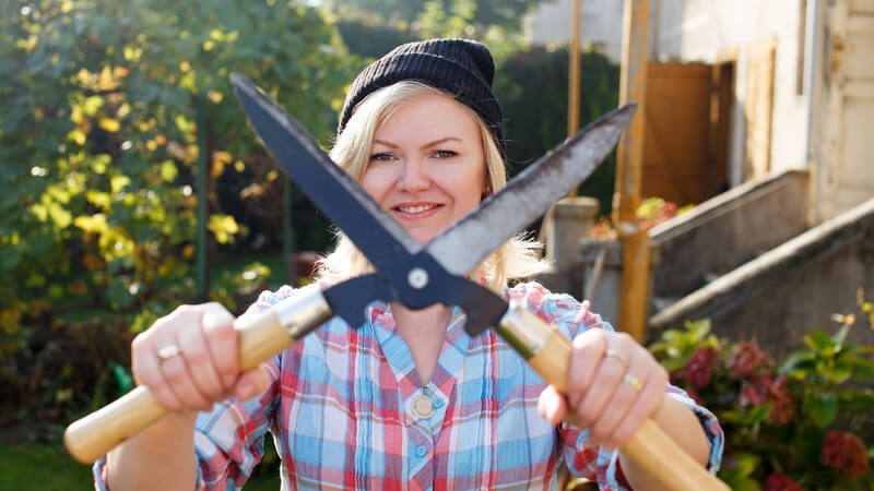 Blonde Frau steht mit Mütze im Garten und hält eine große Heckenschere