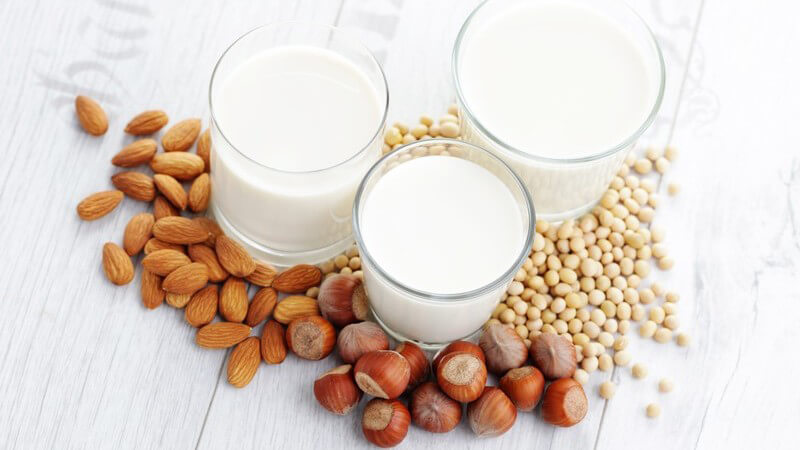 Drei Gläser mit verschiedenen veganen Milchsorten, davor liegen Mandeln, Nüsse und Soja