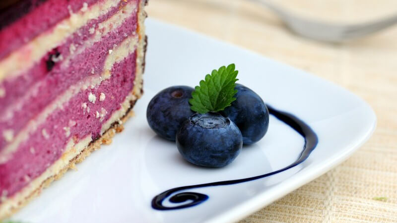 Nahaufnahme Stück Heidelbeertorte, daneben drei Heidelbeeren mit blauer Farbe