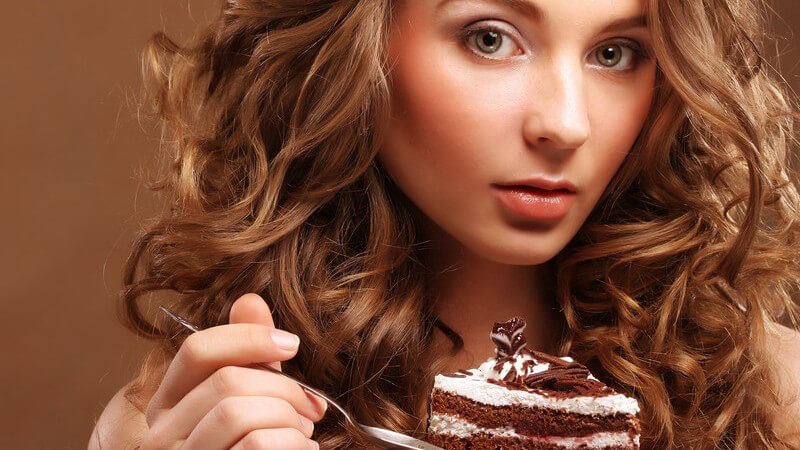 Junge Frau mit langen braunen Locken isst ein Stück Torte vor braunem Hintergrund