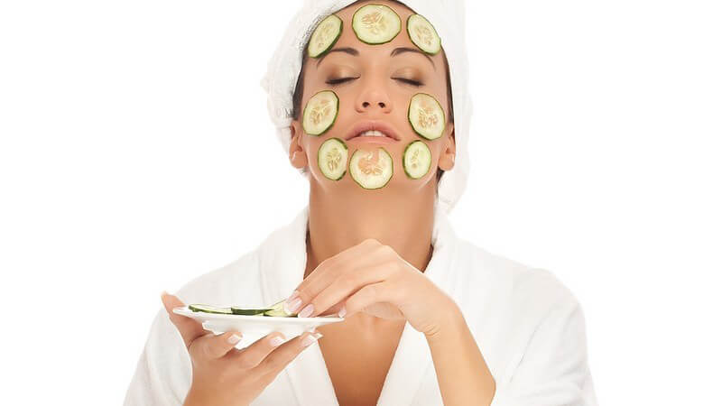 Frau in weißem Bademantel sitzt im Schneidersitz, legt sich Gurkenscheiben auf Gesicht