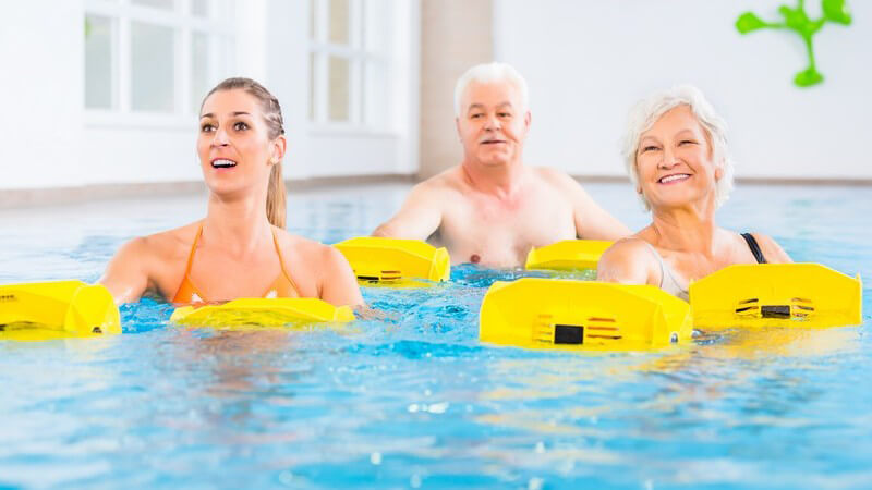 Junge Frau und ein älteres Paar bei der Wassergymnastik mit gelben Hilfsmitteln in den Händen