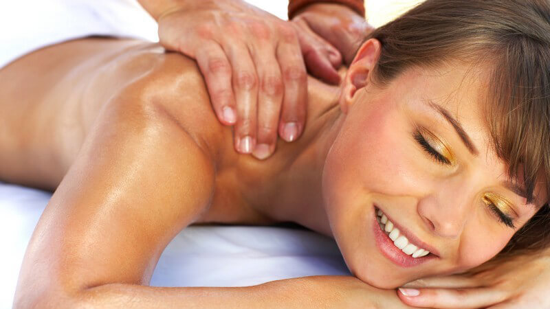 Junge Frau mit nacktem Oberkörper liegt auf dem Bauch und bekommt Schultermassage